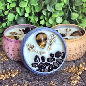 May include: Three round metal candle tins in light blue, pink, and yellow, each featuring a white wax surface topped with a polished tiger's eye stone, two small gold-toned metal keys, and several smooth black obsidian stones.