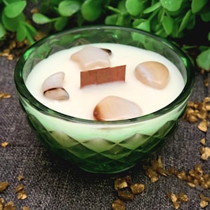 May include: A scented candle in a green glass bowl with a diamond-cut pattern. The white wax contains a rectangular wooden wick and four smooth, polished stones in the shapes of a star, a crescent moon, and two rounded pebbles.