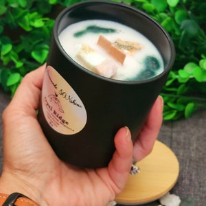 May include: A black candle with a matte finish, featuring a white wax top and decorative wooden pieces. The candle has a label that reads "Homemade & Shine" and "Opal Ridge". The candle is held in a hand, with a wooden lid and a brown leather watch in the foreground.