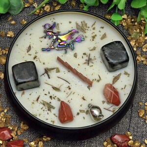 May include: A round black candle with a white wax interior, featuring a metallic witch on a broom, crystals, and herbs. The candle is in a black container. Red and black stones are scattered around.