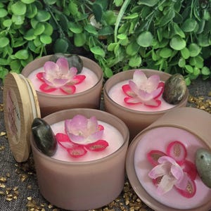 May include: Four pink candles with floral accents and decorative stones. Each candle is in a round, frosted container. The candles have a light pink wax and are topped with a pink and white flower. A wooden lid is visible.