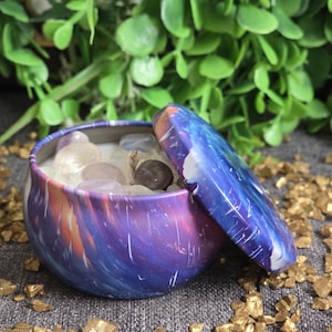 May include: A small, round container with a lid, featuring a swirling galaxy design in shades of purple, blue, and orange. The container is open, revealing a collection of small, polished gemstones. The background includes green foliage and gold-coloured rocks.