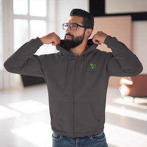 Könnte beinhalten: Anthrazitfarbener Kapuzenpullover mit Reißverschluss und kleinem grünem Logo auf der Brust. Der Hoodie hat einen durchgehenden Reißverschluss, eine Kapuze und zwei Vordertaschen. Das Logo ist ein stilisiertes Design mit Text darunter.