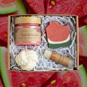 May include: A gift set featuring watermelon-themed bath products. It contains a jar of pink foaming sugar body scrub, a watermelon soap, a white knot candle, and a lip balm. The box is set against a backdrop of watermelon slices.