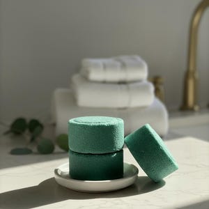 May include: Three teal-colored, cylindrical bath bombs stacked on a small white ceramic dish. One bath bomb is leaning against the others. White towels are stacked in the background, suggesting a spa or bathroom setting.