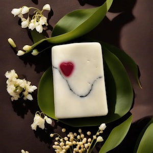 May include: A white bar of soap with a black swirl design and a red heart on a green leaf. The soap is surrounded by white flowers.