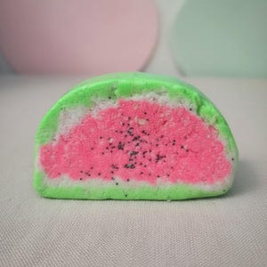 May include: A watermelon-shaped bath bomb with a green rind, white flesh, and pink center dotted with black seeds. The bath bomb is a half-circle shape, and the colors are vibrant and distinct. The background is a neutral color.