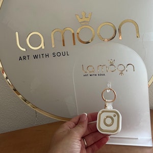 May include: A white and gold keychain with a camera design. The keychain is on a white display stand with the text "la moon art with soul" on it.