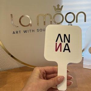 Puede incluir: Espejo de mano blanco con un logotipo "ANNA" negro y rosa. El espejo se sostiene en una mano frente a un letrero redondo dorado con el texto "la moon ART WITH SOUL" y una corona.