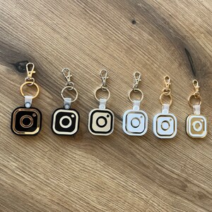 May include: Seven keychains with different colored Instagram logo charms. The charms are gold, black, white, and silver. The keychains are all attached to silver rings.