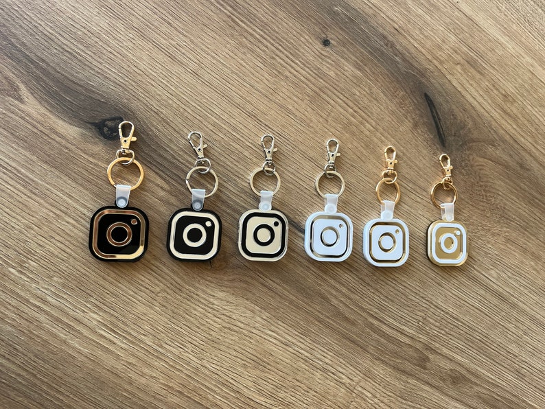 May include: Seven keychains with different colored Instagram logo charms. The charms are gold, black, white, and silver. The keychains are all attached to silver rings.