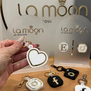 May include: A collection of keychains featuring various designs, including a heart, a camera, a letter E, and the letter F. The keychains are made of white and black plastic with gold accents. The keychains are displayed on a white surface with the brand name "La moon Art with Soul" printed on the surface.