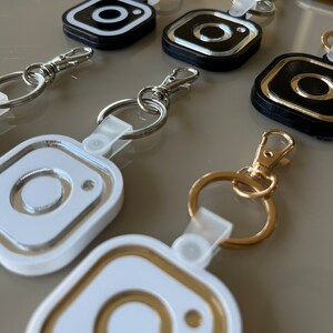 May include: A set of five keychains featuring the Instagram logo. The keychains are made of white, black, and gold plastic. Each keychain has a different color combination.