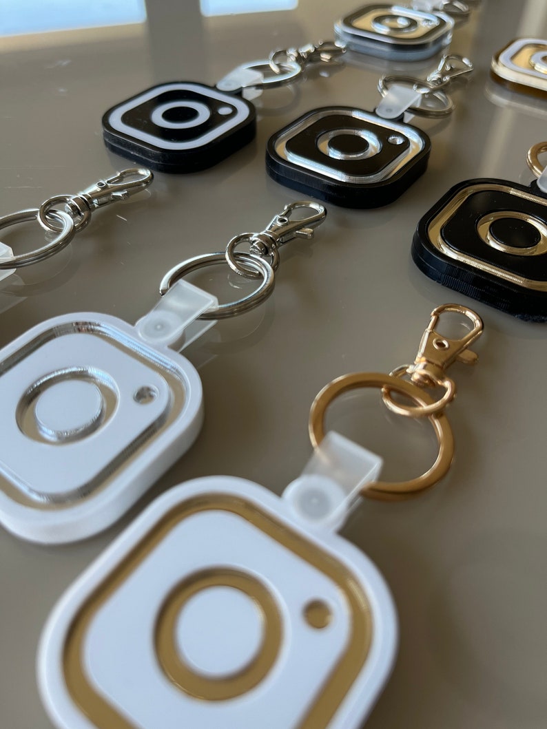 May include: A set of five keychains featuring the Instagram logo. The keychains are made of white, black, and gold plastic. Each keychain has a different color combination.