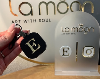 May include: Black leather keychain with a gold letter E. The keychain is attached to a silver key ring. The keychain is displayed on a white stand with the text "la moon art with soul".
