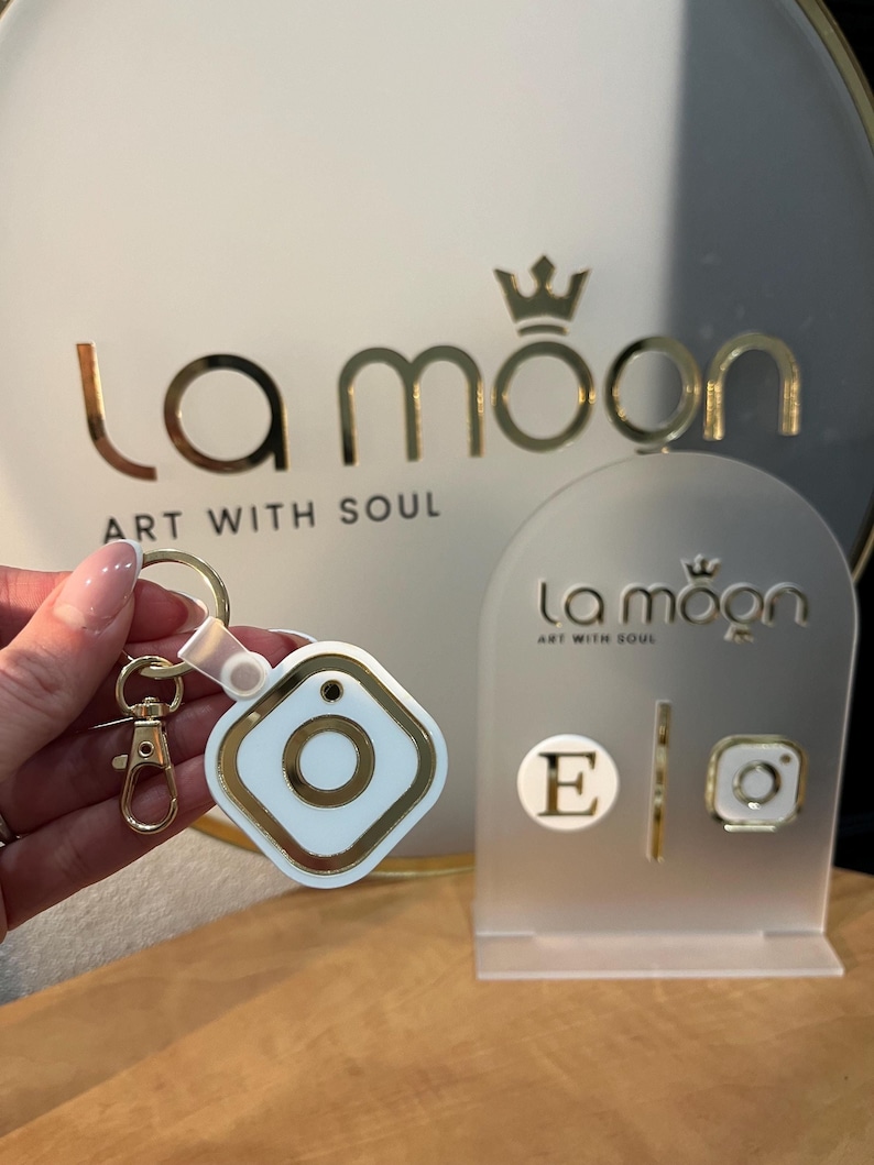 May include: A white and gold keychain with a square charm featuring a gold circle inside. The charm is attached to a gold chain and a gold split ring. The keychain is displayed on a white stand with the text "la moon art with soul" in gold lettering.