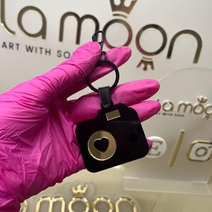 May include: Black camera-shaped keyring with a gold heart cutout. The keyring is attached to a black metal ring and a silver chain.