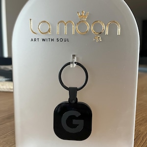 May include: Black keyring with a square charm featuring a silver letter G on a black background. The keyring is displayed on a white stand with the text "la moon ART WITH SOUL" in gold lettering.