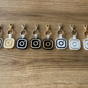 May include: Eight keychains with different colored Instagram logo charms. The charms are black, white, gold, and silver. The keychains are silver.