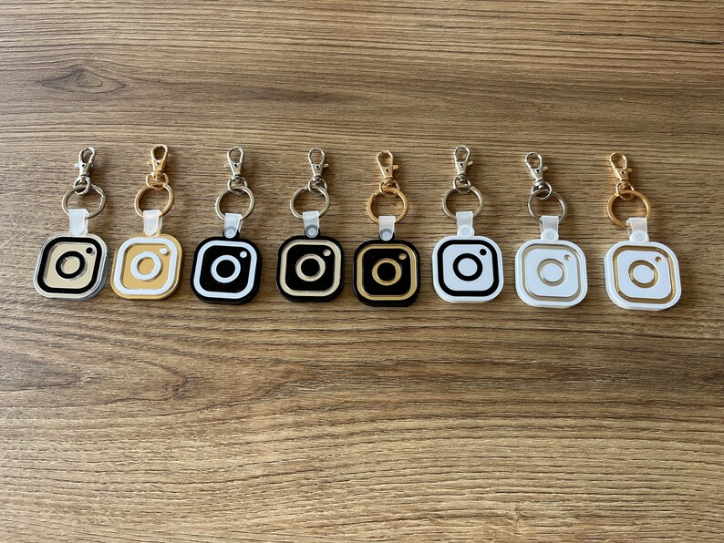 May include: Eight keychains with different colored Instagram logo charms. The charms are black, white, gold, and silver. The keychains are silver.