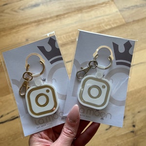 May include: Two white keychains with gold accents shaped like an Instagram logo. The keychains are packaged on white cards with the text "la moon art with soul".
