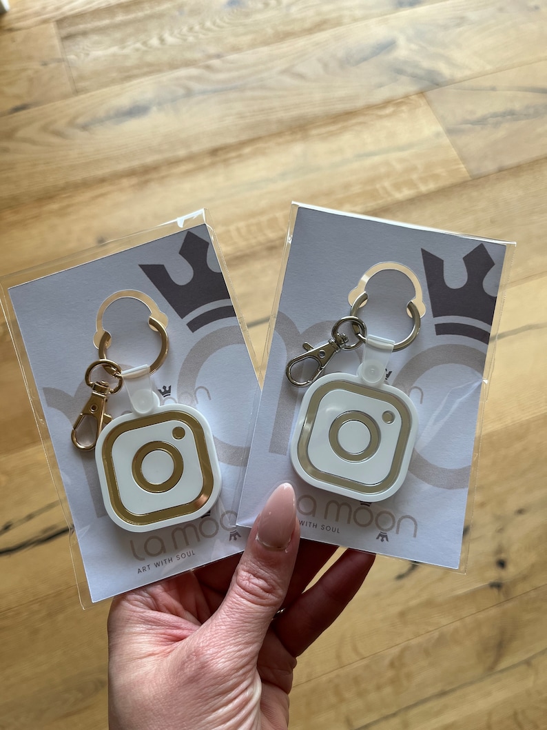 May include: Two white keychains with gold accents shaped like an Instagram logo. The keychains are packaged on white cards with the text "la moon art with soul".
