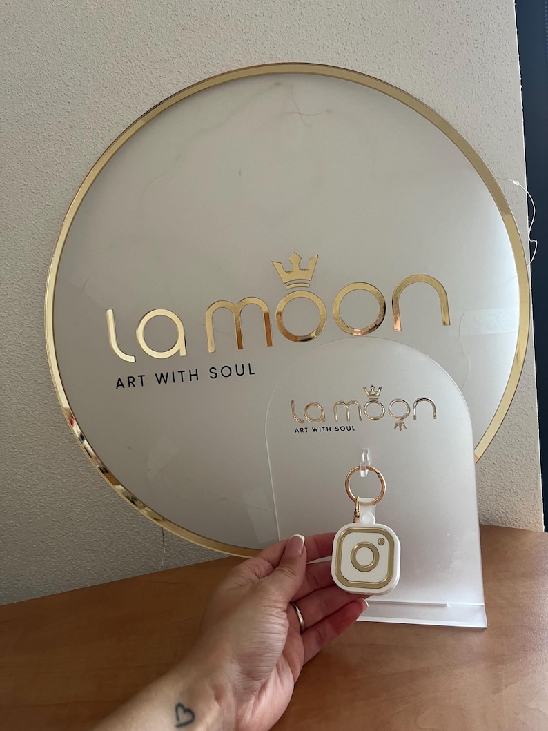 May include: A white and gold keychain with a camera design. The keychain is on a white display stand with the text "la moon art with soul" on it.