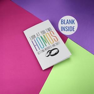 May include: A white greeting card with the text "LOOK AT YOU TWO HOMOS GETTING MARRIED AND SHIT!" in rainbow colours. Two black wedding rings are below the text. A white circle with "BLANK INSIDE" is in the upper right corner.