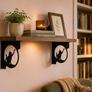 May include: A wooden wall shelf supported by two black metal brackets, each featuring a circular cutout with a silhouette of a cat reaching for a butterfly. The shelf holds a potted plant, a lit candle in a glass jar, and a framed botanical print.