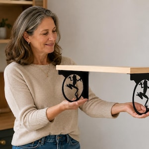 May include: A wall-mounted shelf featuring a light-colored wooden plank supported by two black metal brackets. Each bracket has a circular design containing a silhouette of a frog in a jumping position, providing a decorative and functional support.