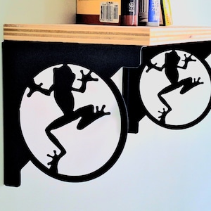 May include: Two black metal shelf brackets with a frog silhouette design. Each bracket features a circular cutout with a white background and a black frog silhouette.