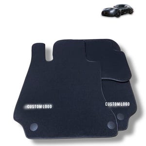 Puede incluir: Un juego de alfombrillas negras para coche con superficie texturizada y bordes oscuros. Cada alfombrilla tiene una fijación circular en la parte inferior y el texto CUSTOM LOGO impreso en blanco. Una pequeña imagen de un coche deportivo negro arriba.