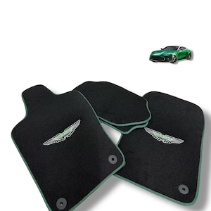 May include: Black car floor mats with green trim and the Aston Martin logo embroidered in white and green. A green sports car is in the background.