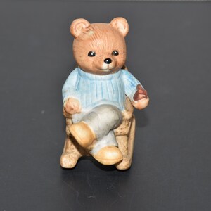 May include: A brown ceramic teddy bear wearing a blue sweater sits in a rocking chair. The bear is holding a small brown object in its right paw.