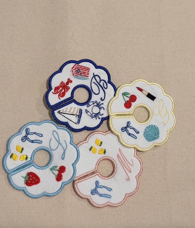 May include: Embroidered white fabric coasters with scalloped edges. Each coaster features a unique design, including a lobster, sailboat, cherries, lemons, and initials. The coasters have a hole in the center.