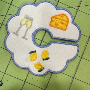 May include: White fabric coaster with a blue scalloped edge, embroidered with champagne glasses, a cheese wedge, and lemons. The coaster has a circular cutout in the center.