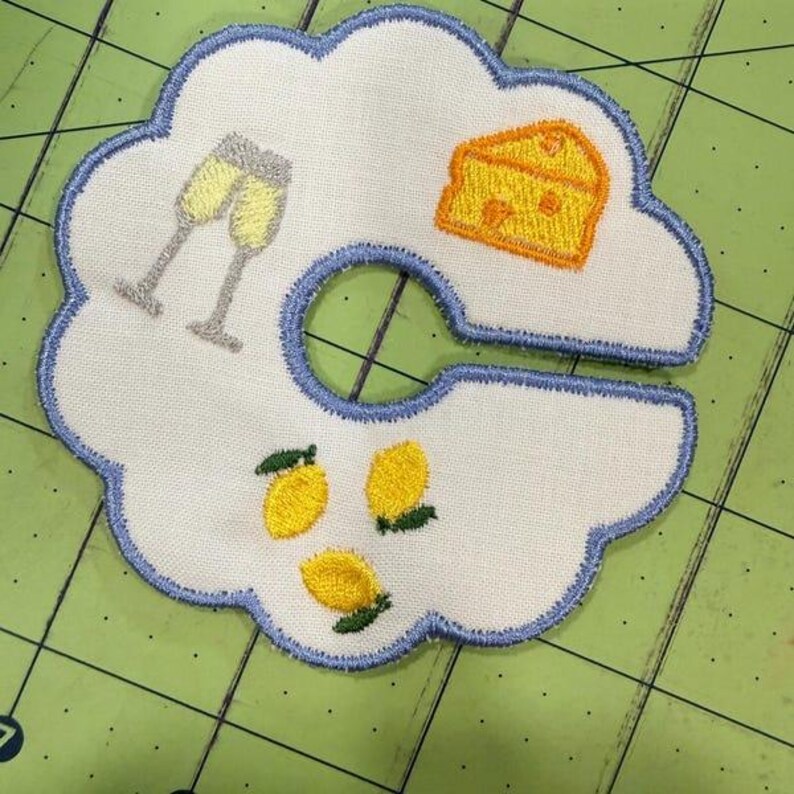 May include: White fabric coaster with a blue scalloped edge, embroidered with champagne glasses, a cheese wedge, and lemons. The coaster has a circular cutout in the center.