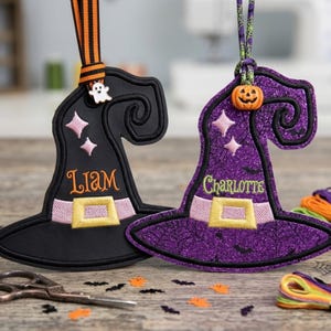 May include: Two witch hat ornaments featuring embroidered details. The black hat has the name Liam, a ghost charm, and orange-striped ribbon. The purple glitter hat has the name Charlotte, a pumpkin bead, and a green spiderweb-patterned ribbon.