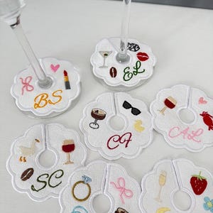 May include: White embroidered wine glass charms with various designs, including a lipstick, coffee bean, martini, and a ring. Each charm has a unique initial. These charms are perfect for a party or event.