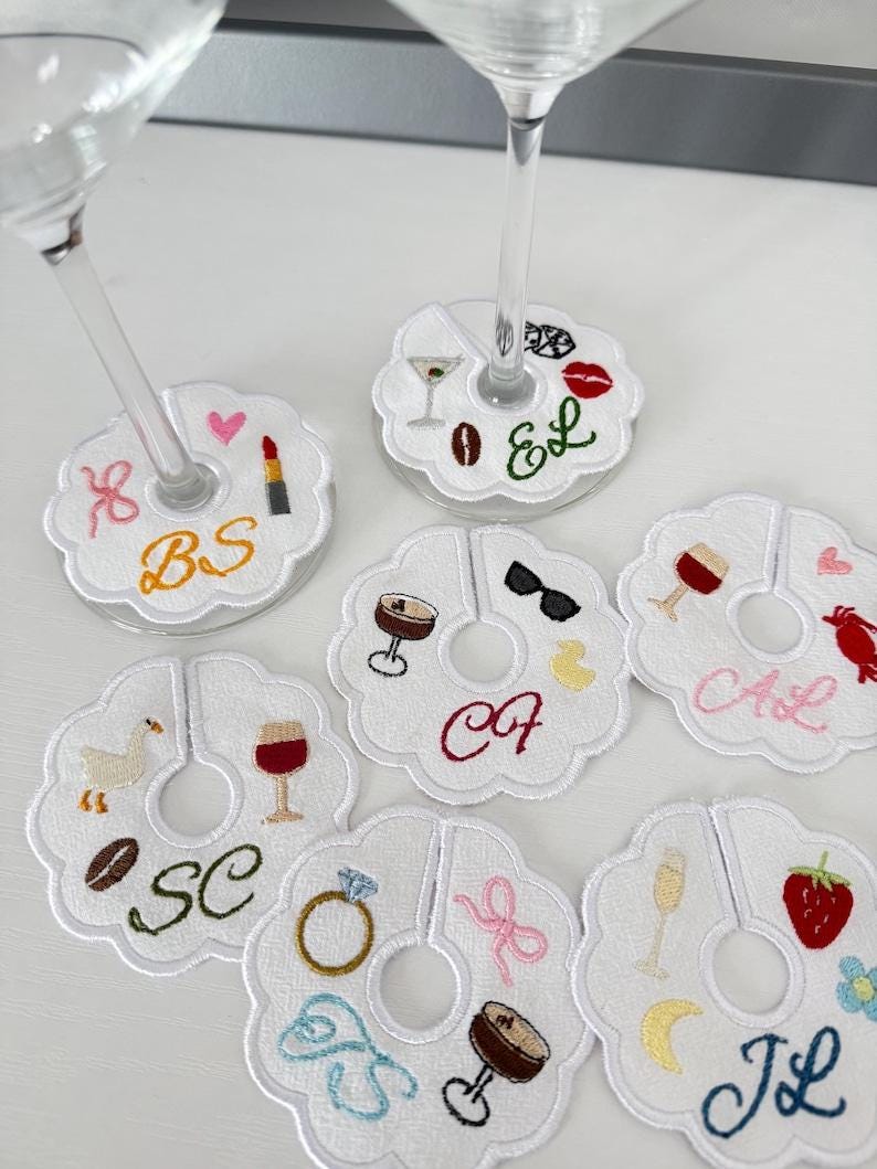 May include: White embroidered wine glass charms with various designs, including a lipstick, coffee bean, martini, and a ring. Each charm has a unique initial. These charms are perfect for a party or event.