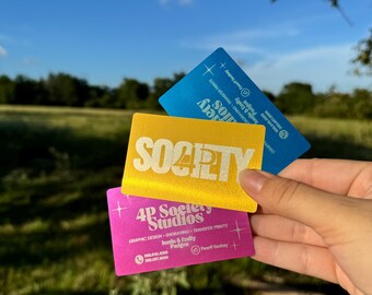 May include: Three business cards with a gold, pink, and blue background. The gold card has the text "Society" in a retro font. The pink card has the text "4P Society Studios" and the text "Graphic Design, Engraving, Transfer Prints." The blue card has the text "Society" and the text "Emily & Ashley."
