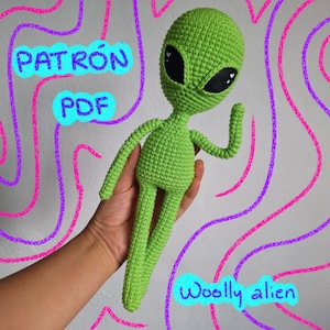 Puede incluir: Un extraterrestre de crochet verde con ojos grandes y un diseño en forma de corazón en cada ojo. El extraterrestre está levantando su brazo derecho en señal de saludo. El texto "PATRÓN PDF" está en la esquina superior izquierda de la imagen. El texto "Woolly alien" está en la esquina inferior derecha de la imagen.