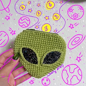 May include: A green crocheted alien-shaped coin purse with black sequined eyes and a green zipper. The words "Woolly alien" are written on the bottom right. The background features doodles of planets, stars, and dollar signs.