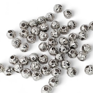 May include: A collection of small, silver-colored beads with a textured, hammered appearance. The beads are scattered on a white surface, each with a hole for stringing. These beads are suitable for jewelry making and crafts.