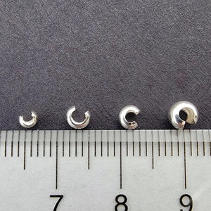 May include: Four silver crimp beads of different sizes, measured on a ruler in inches. The beads are 6/8, 7/8, 8/8, and 9/8 inches in diameter.