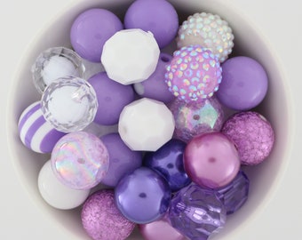 May include: A bowl filled with a variety of purple, white, and iridescent beads. The beads are made of different materials, including plastic, glass, and metal. Some of the beads have a shiny finish, while others have a matte finish.