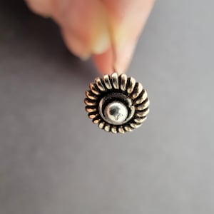 May include: A silver nose ring with a decorative flower design. The flower has a center circle with a raised, textured surface.