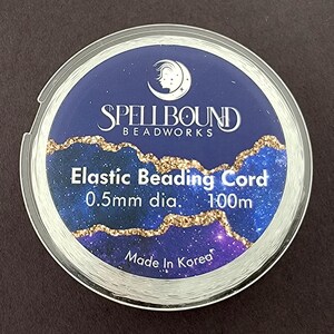 K&ouml;nnte beinhalten: Eine Spule mit transparentem elastischem Schmuckband. Das Etikett ist dunkelblau mit goldenen Glitzerakzenten und dem Text "Spellbound Beadworks". Der Text "Elastic Beading Cord" und "0,5 mm Durchmesser. 100 m" sind ebenfalls sichtbar. Unten steht "Made in Korea".