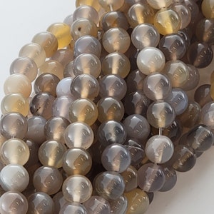 May include: Close-up of a strand of round, polished beads in shades of gray, brown, and yellow. The beads are strung together, creating a textured surface. The beads are approximately 8mm in diameter.