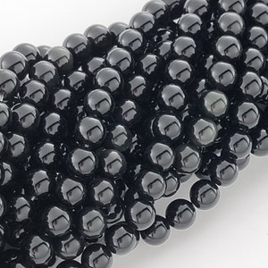 May include: A close-up of a strand of black gemstone beads. The beads are round and smooth, with a high polish. They are arranged in a random pattern, creating a visually appealing texture.